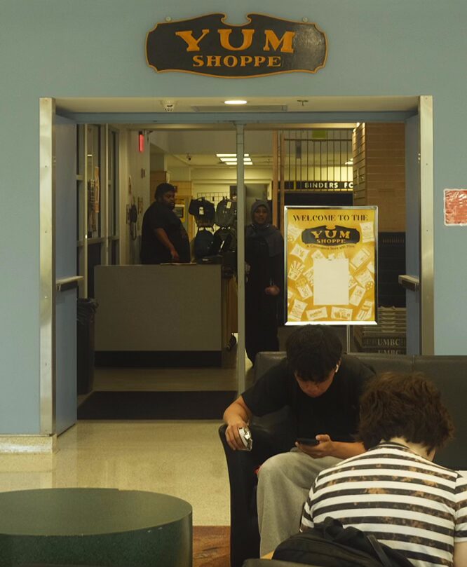Photo of the Yum Shoppe downstairs entrance, with the double doors open and a background of students going about their lives and purchasing things from the Yum Shoppe checkout.